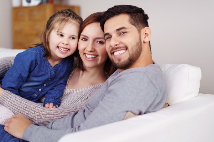 Familie auf einer Couch, lächelnd, mit einem Kind zwischen den Eltern.