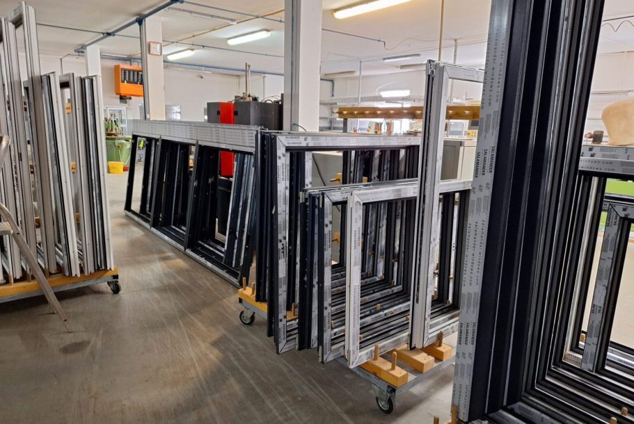 Enovia Haus Fensterwerk Eine Reihe von Rahmen in verschiedenen Größen und Farben in einer Fabrikhalle.