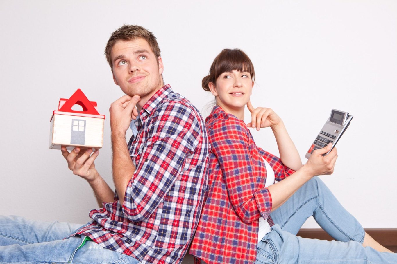 Junges Paar sitzt Rücken an Rücken, denkt nach, hält ein Hausmodell und ein Smartphone.