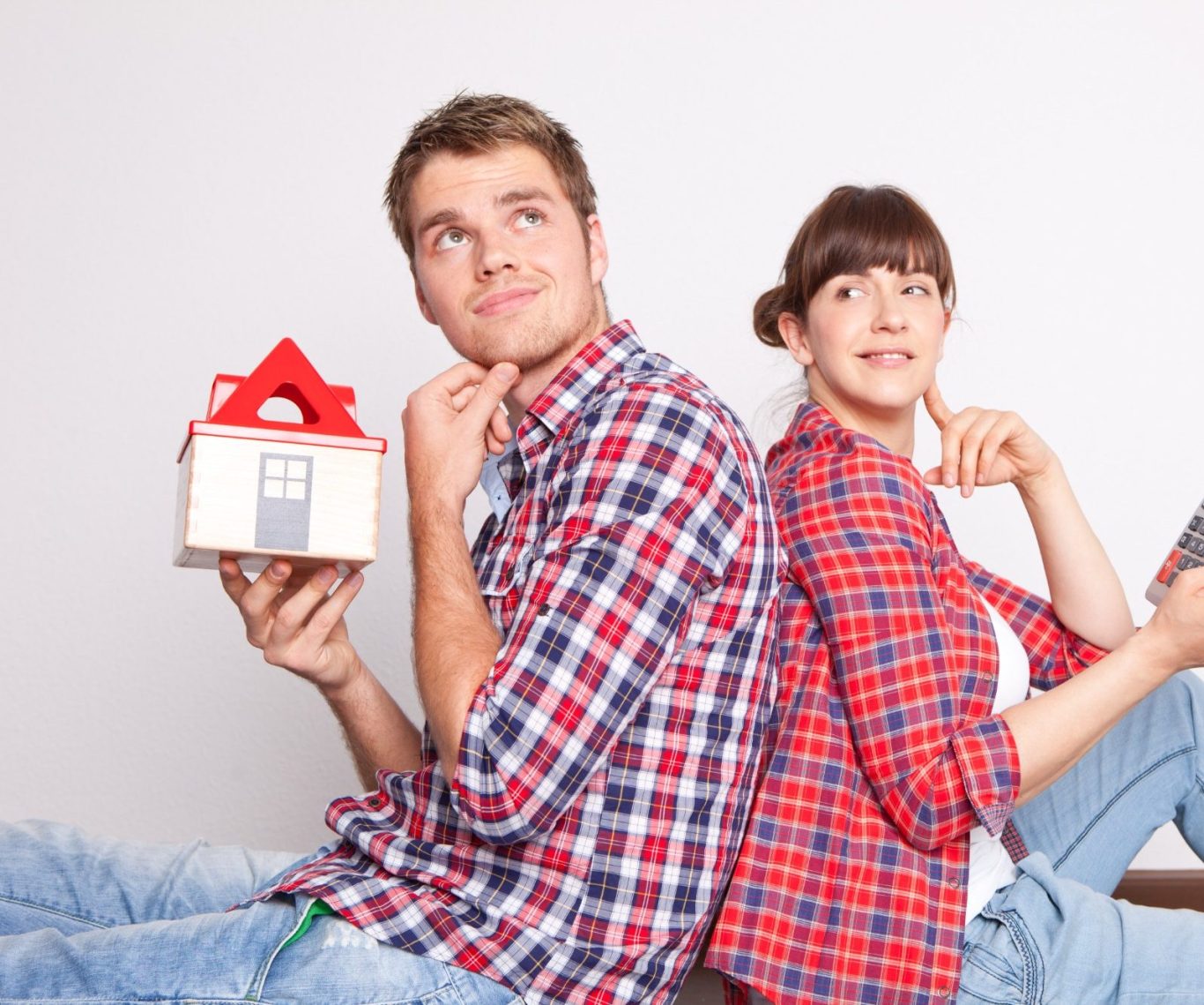 Junges Paar sitzt Rücken an Rücken, denkt nach, hält ein Hausmodell und ein Smartphone.