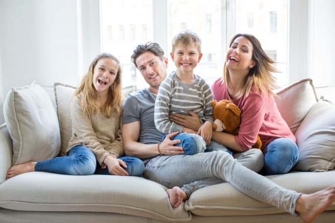 Eine fröhliche Familie sitzt zusammen auf einem Sofa und lächelt in die Kamera.