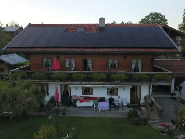 Ein zweigeschossiges Holzhaus mit Balkon, umgeben von Grünflächen und mehreren Personen.