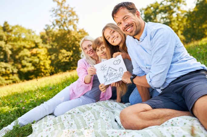 Eine Familie sitzt auf einer Decke im Freien und hält eine Zeichnung hoch.