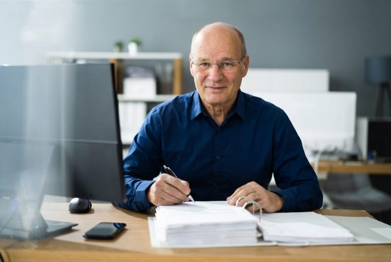 Älterer Mann in Hemd sitzt am Schreibtisch und notiert etwas. Computer im Hintergrund.