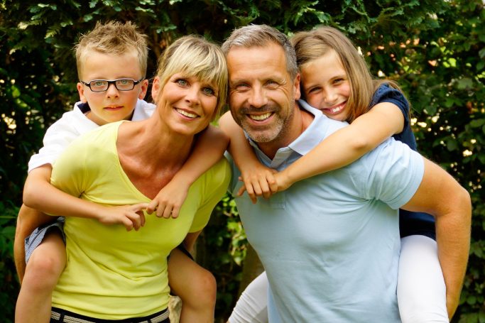 Familie mit zwei Kindern, das Paar lächelt, die Kinder sitzen auf den Schultern der Eltern.