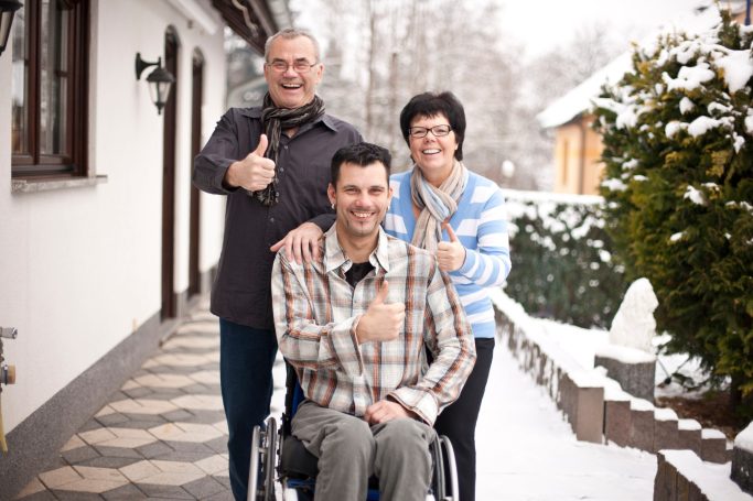Drei Personen, ein Mann im Rollstuhl, posieren lächelnd vor einem Haus im Schnee.